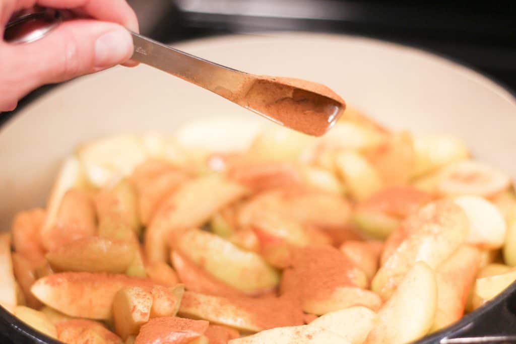A measuring spoon pouring ground cinnamon over apples cooking in a pot.