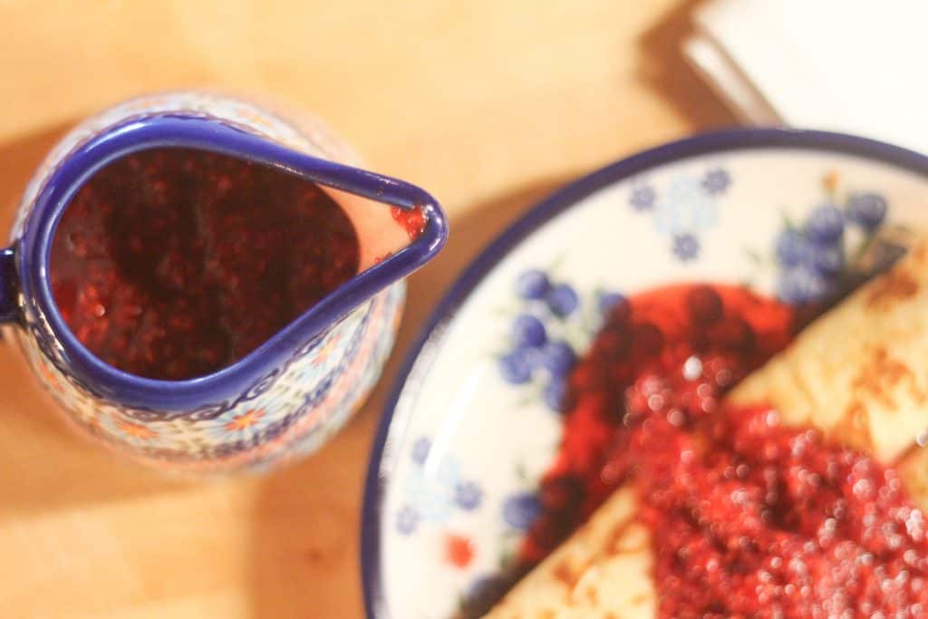 A pitcher of berry sauce by a plate of crepes