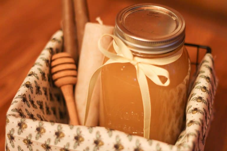 Inside a black basket lined with bee fabric and filled with a jar of honey, a wooden honey dipper, and two candles