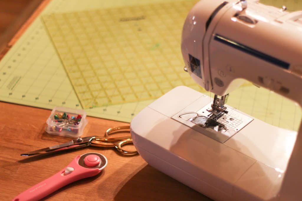 A sewing machine, rotary cutter, scissors, pins, and a cutting mat. Materials to make a diy potholder