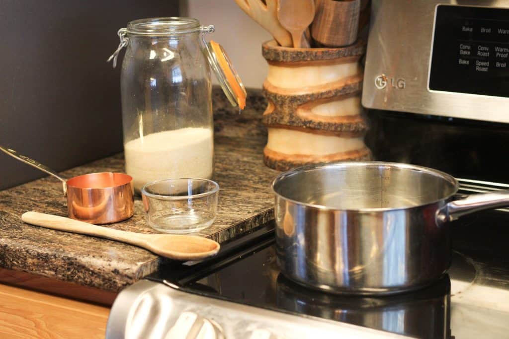 A stovetop with a saucepan on it and a jar of sugar, a measuring cup, and a wooden spoon sitting on the counter