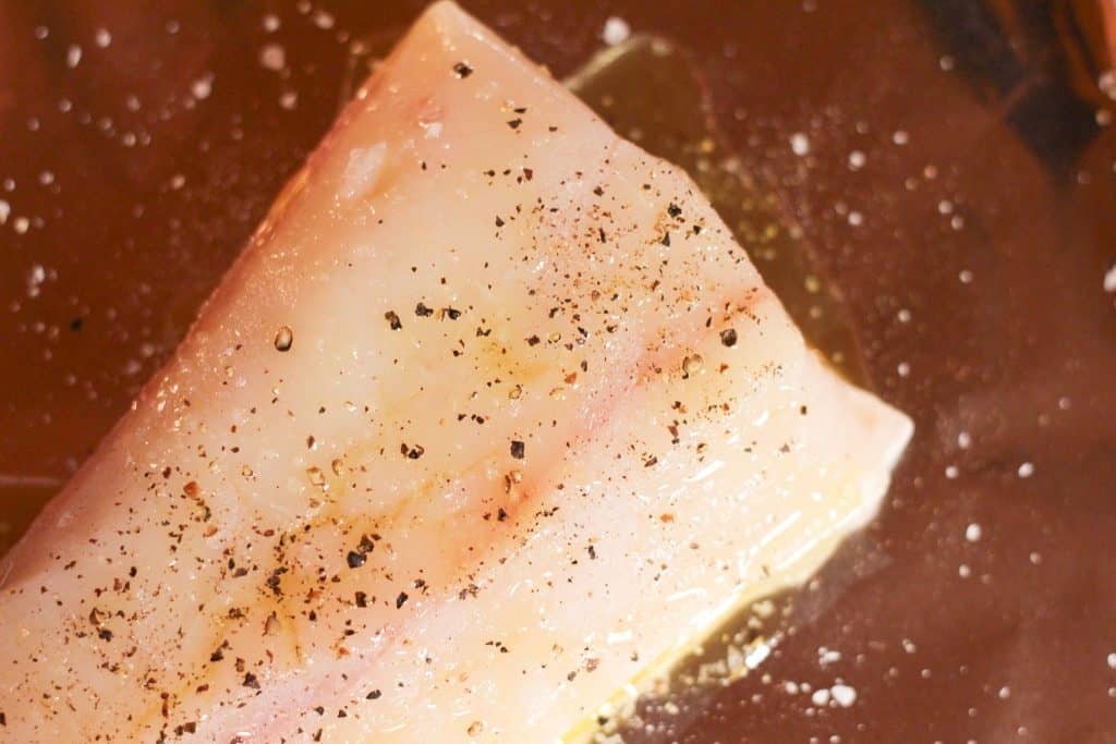 A piece of raw white fish that has been sprinkled with salt, pepper, and olive oil
