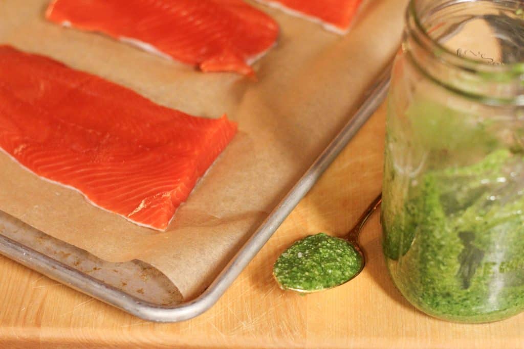 A spoonful of pesto next to a glass jar of pesto sauce and some salmon fillets on a baking pan